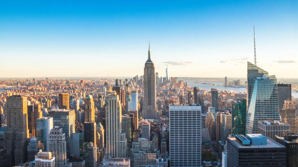 Ein Panoramablick auf die Skyline von New York City bei Sonnenuntergang mit hohen Wolkenkratzern, darunter das Empire State Building, mit klarem blauen Himmel und im Hintergrund sind in der Ferne Flüsse zu sehen.